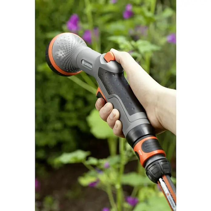 Gardena Comfort Brause - Anzahl Sprühformen: 2 Stk., Metall/Kunststoff - Gardena Multifunktions-Brause liegt in einer Hand vor einem Blumenbeet. Der Brausekopf ist grau mit orangenen Akzenten.