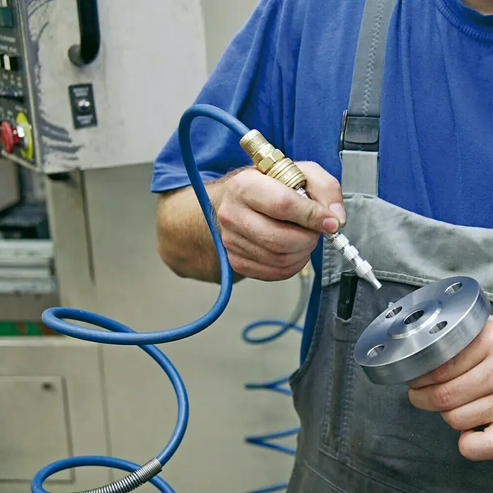 Hazet Ausblasstift 9040-3 - Betriebsdruck: 6 bar, Durchflussmenge: 360 l/min - Ein Arbeiter reinigt ein rundes Metallteil mit einem Druckluftwerkzeug. Der blaue Schlauch ist an die Werkzeugmaschine angeschlossen.