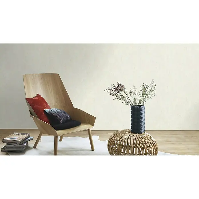 Rasch Vliestapete - Creme, Uni, 10,05 x 0,53 m, Spachtelstruktur - Eine entspannte Interieur-Szene mit einem hellen Holzstuhl, Kissen, Büchern und einer Vase mit Blumen auf einem runden Beistelltisch.