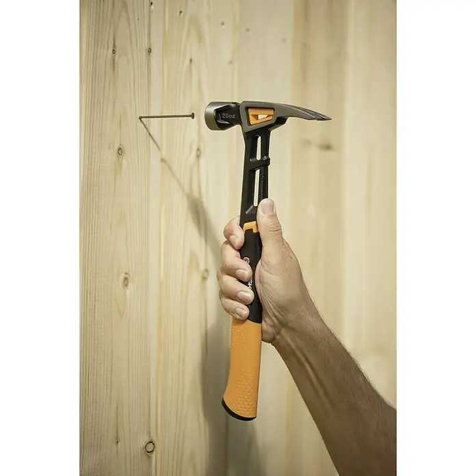 Fiskars Universalhammer M - Gewicht Kopf: 450 g, Länge: 340 mm - Nahaufnahme eines Mannes, der einen Fiskars Hammer benutzt, um einen Nagel in eine Holzwand zu schlagen. Der Hammer hat einen orangefarbenen Griff.