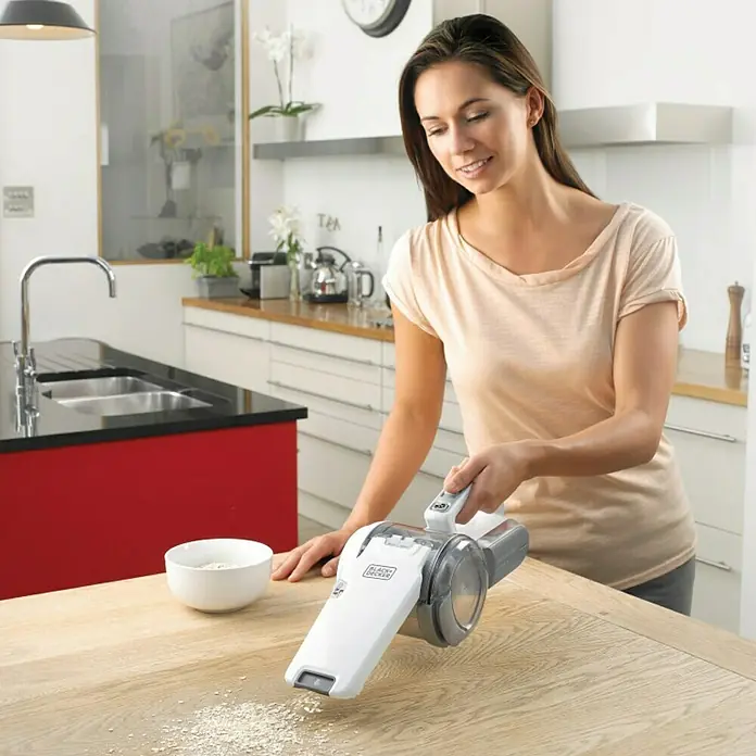 Black+Decker Aspirador de mano de batería PV1820L-AARU - 18 V, 1 batería, Volumen de recipiente: 440 ml - Una mujer está utilizando una aspiradora de mano Black+Decker para limpiar migas de una mesa de madera en una cocina moderna.