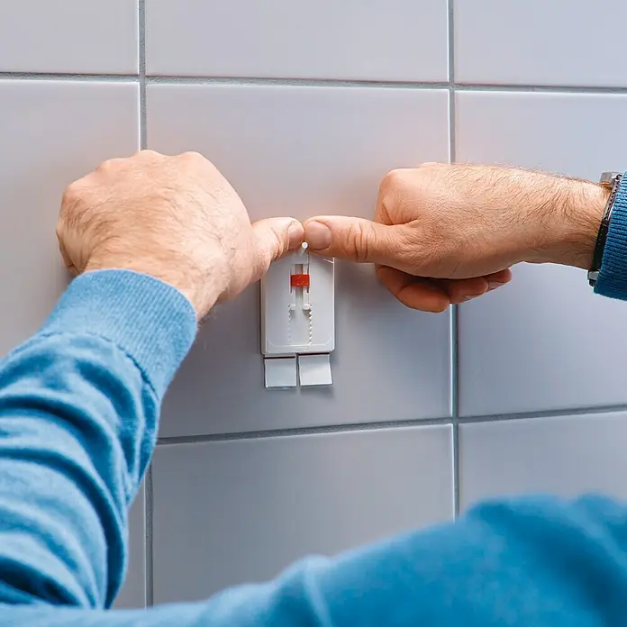Tesa Clavo adhesivo ajustable - Blanco, Carga soportada: 4 kg, 2 ud. - Un hombre está pegando un gancho adhesivo blanco con detalles rojos a una pared de azulejos. El gancho adhesivo es adecuado para baños o cocinas.