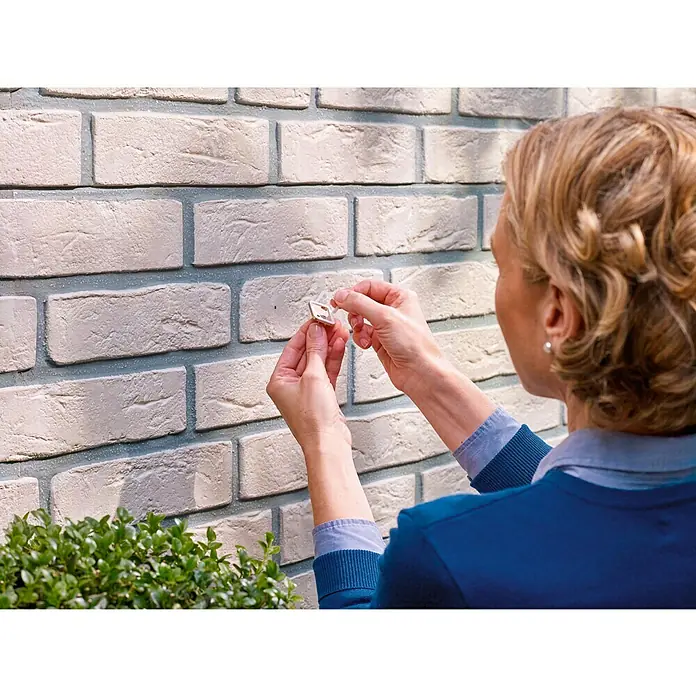 Tesa Tornillo adhesivo - Rectangular, Carga soportada: 5 kg, 2 ud. - Mujer que sujeta un gancho adhesivo a una pared de ladrillos. El gancho es discreto y fácil de aplicar.
