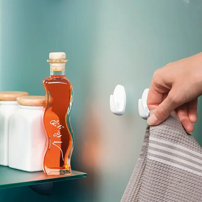 Tesa Powerstrips Gancho autoadhesivo Mini Gancho Ovalado - Blanco, 3 ud. - Una naturaleza muerta de baño que muestra dos ganchos de toalla blancos en una pared tonalizada, una toalla y botes decorativos en un estante de vidrio.