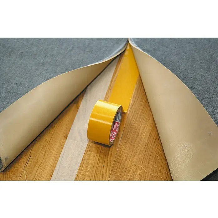 Tesa Cinta de doble cara Universal Moquetas - 50 mm x 10 m, Beige - Un rollo de cinta de doble cara Tesa entre dos trozos de moqueta sobre un suelo de madera. Ideal para fijar moquetas.