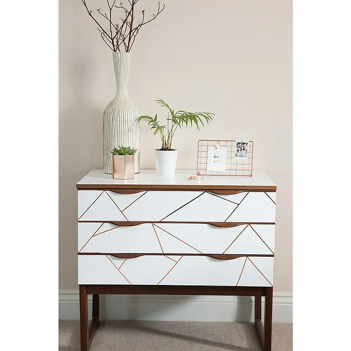 D-c-fix Klebefolie Uni Seidenmatt - 200 x 45 cm, Weiß, Selbstklebend - Weiße Kommode mit geometrischen Kupferlinien und Holzgriffen, dekoriert mit Pflanzen, Vase und Fotorahmen.