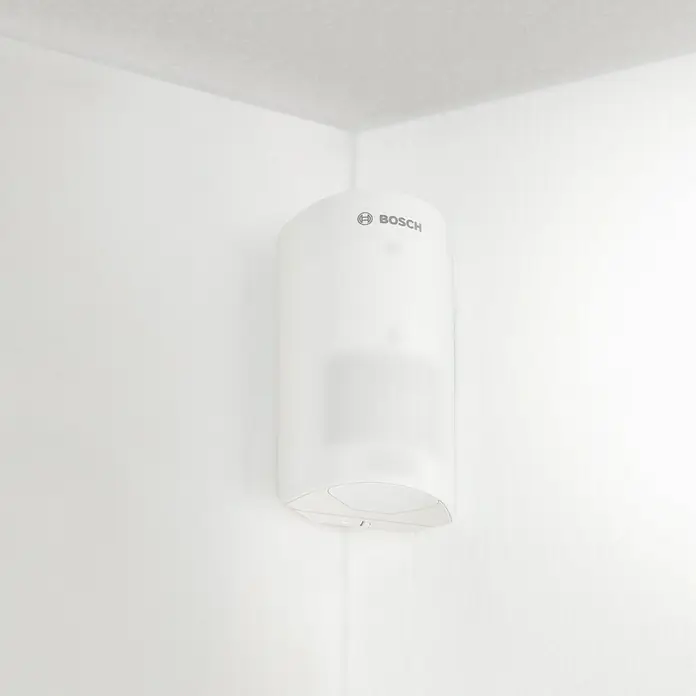 Bosch Smart Home Bewegungsmelder - Smarte Steuerung: Bosch Smart Home App, Reichweite Funk: 50 m - Nahaufnahme eines weißen Bosch-Geräts, das an einer weißen Wand in einer Ecke des Raumes montiert ist.
