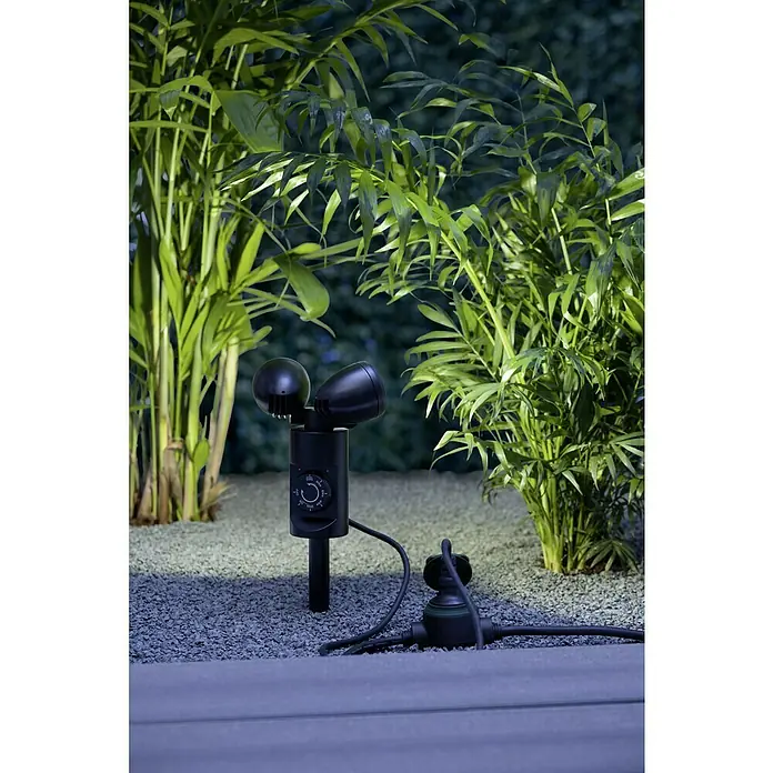 Ritter Leuchten LED-Gartenspot - 6 W, Höhe: 140 mm, Erdspieß, Kunststoff - Gartenbeleuchtung mit zwei Scheinwerfern und einem Verteiler in einem Kiesbeet, umgeben von üppigen Pflanzen.