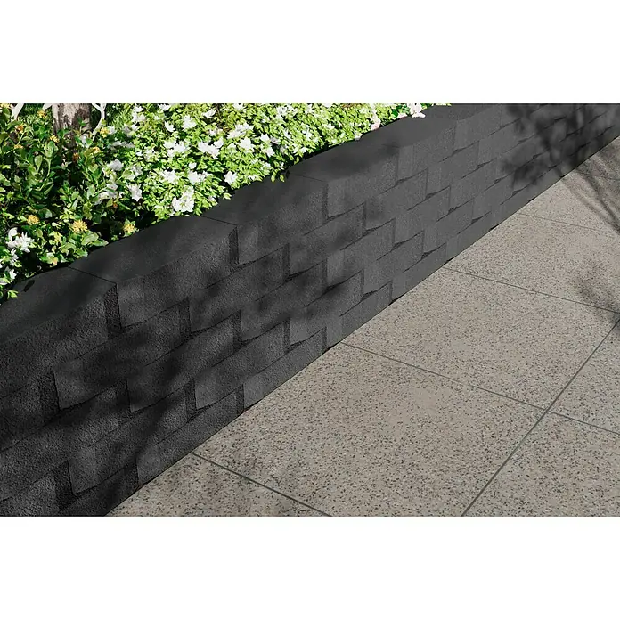 Christoph Mauerstein Antikblock - 30 x 15 x 7,5 cm, Anthrazit, Beton - Nahaufnahme einer stilvollen Mauer aus strukturierten schwarzen Steinen neben einem gepflasterten Bereich und üppigem Grün.