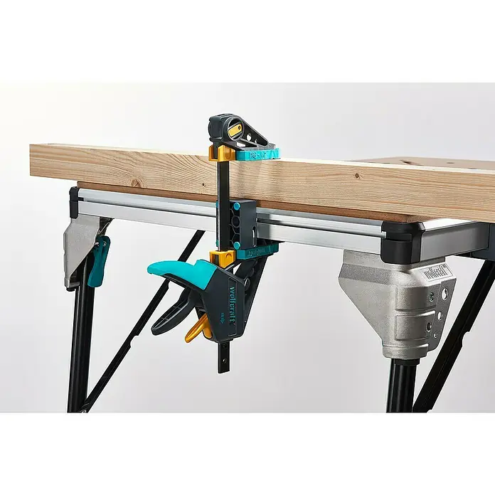 Wolfcraft Werkbank Master Work 1600 - L x B: 76,5 x 110 cm, Arbeitshöhe: 86,3 cm, Belastbarkeit: 120 kg - Eine Wolfcraft Einhandzwinge fixiert ein Holzbrett auf einer Werkbank. Die Zwinge ist blau-schwarz und hält das Brett sicher fest.