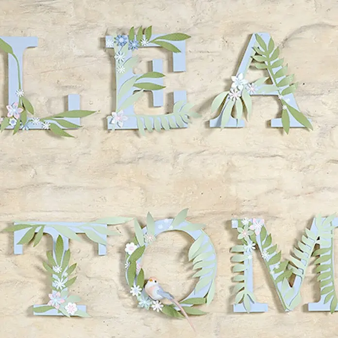 Artemio Letra - 1,3 cm, Madera, Natural/marrón claro, 16 ud. - Letras decorativas "LEATOM" con elementos florales y un pajarito sobre una pared de piedra.