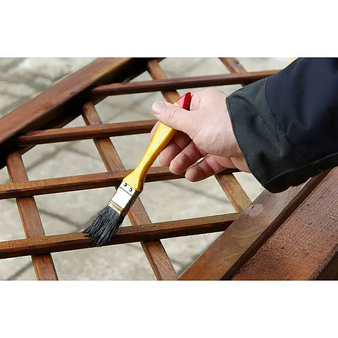 Promade Protección para madera Lasur al agua - Nogal, 750 ml, Mate - Un hombre pinta una celosía de madera con un pincel amarillo. La madera se oscurece y la veta se hace visible.