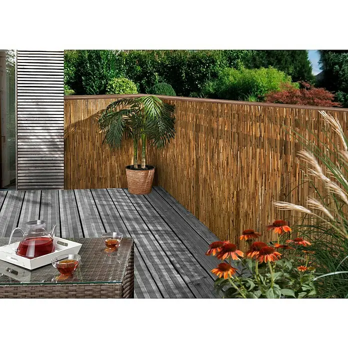 Gardol Sichtschutzmatte - 3 x 1 m, Schilfrohr - Ein entspannender Außenbereich mit Bambuszaun, komfortablen Terrassendielen, einem Tisch mit Getränken und blühenden Blumen.