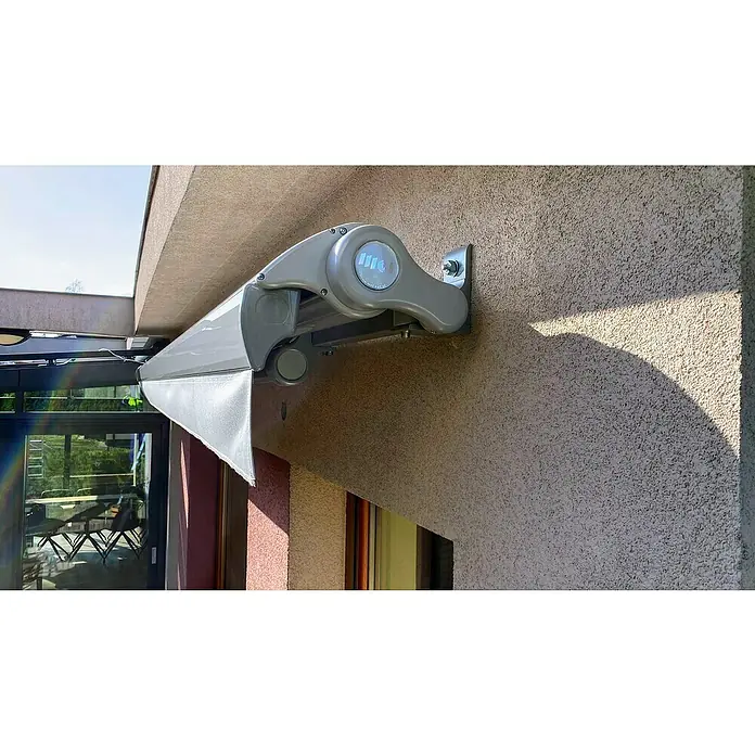 SM Hülsenmarkise REC-196 - Hellgrau, Farbe Gestell: Weißaluminium, Breite: 5 m, Ausfall: 3 m - Nahaufnahme einer eleganten, grauen Markise, die an einer getexturieren Wand montiert ist und Schatten spendet.\n