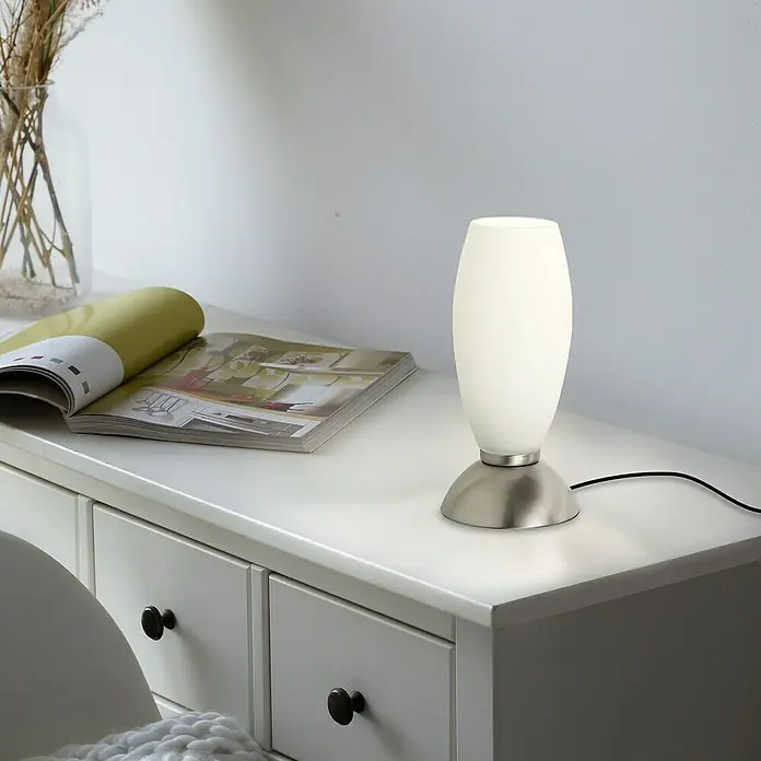 Paul Neuhaus LED-Tischleuchte Joy - L x B x H: 8,5 x 8,5 x 20 cm, Stahl, Warmweiß - Elegante weiße Tischleuchte mit silbernem Sockel auf einer Kommode, neben einer Zeitschrift und einer Vase mit Pflanzen.