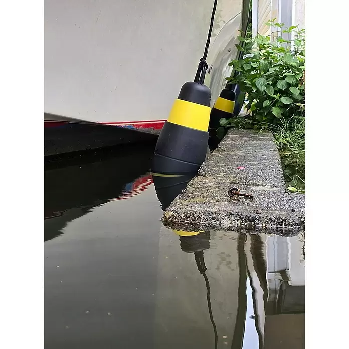 Defensa multifender Zebra - Diámetro: 16,5 cm, Ajustable: 25 cm - 62 cm, Blanco/Negro - Defensas de barco negras y amarillas colgando de un barco, reflejadas en agua tranquila junto a un muelle de hormigón.