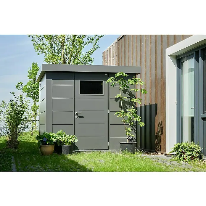 Telluria Metallgerätehaus Classico 2424 - Außenmaß inkl. Dachüberstand (B x T): 258 x 258 cm, Stahl, Anthrazit, Ohne Set - Ein graues Gartenhaus aus Metall steht im Garten. Daneben befinden sich Pflanzen in Töpfen und ein modernes Haus mit Holzfassade.