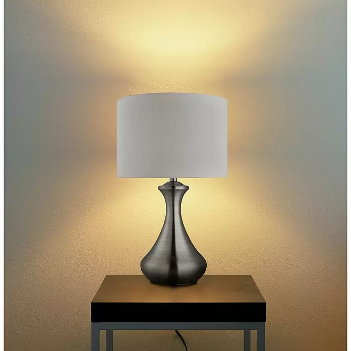 Searchlight Tischleuchte rund Touch - 60 W, 24 x 24 x 40 cm, Silber, E14 - Gebürstete Metall-Tischlampe mit weißem Stoffschirm auf einem Holztisch, beleuchtet eine strukturierte Wand.