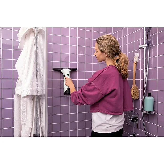 Kärcher Limpiadora de cristales de batería WV 2 Plus N - 1 batería, Volumen de recipiente: 0,1 l - Una mujer usa un aspirador de ventanas Kärcher para limpiar una pared de ducha con azulejos morados, garantizando una limpieza sin rayas.