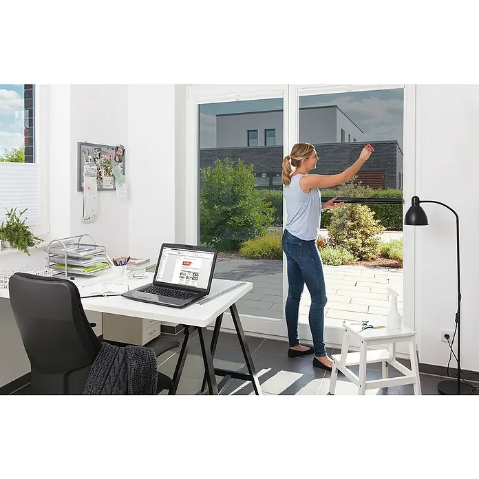 D-c-fix Tönungsfolie - 200 x 90 cm, Mit UV-Schutz, Statisch haftend - Frau reinigt Fenster mit Fensterfolie, um Privatsphäre und Sonnenschutz zu erhöhen. Im Hintergrund ist ein Schreibtisch mit Laptop zu sehen.