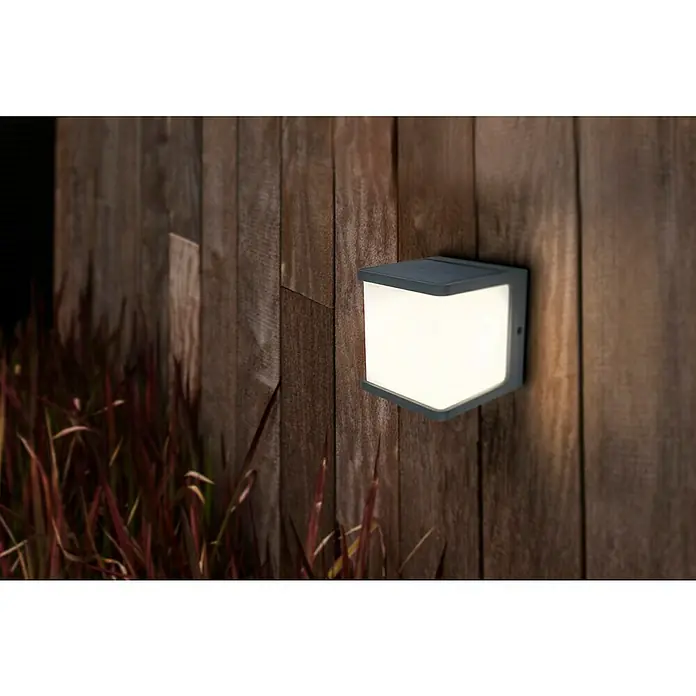 Lutec Doblo Solar-Außenwandleuchte - 7,5 W, L x B x H: 12,9 x 11 x 11 cm, IP54, 500 lm - Quadratische Wandleuchte mit sanftem Licht, montiert an einer rustikalen Holzwand, die einladende Atmosphäre im Außenbereich schafft.