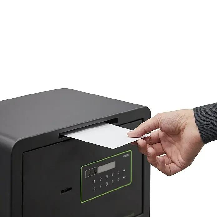 Arregui Kuro Caja fuerte - L x An x Al: 25 x 35 x 25 cm, Negro/Verde, 16 l, Ranura - Una mano está deslizando un sobre blanco en una caja fuerte de seguridad negra con un teclado digital.