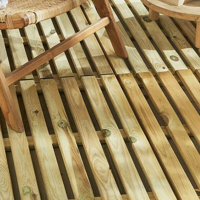 Cerland Loseta de madera Cleia - Pino, 50 x 50 x 3 cm, 1 ud. - Primer plano de un suelo de patio de madera con una pata de silla y una mesa pequeña. Se ven claramente la veta de la madera y los tonos cálidos.