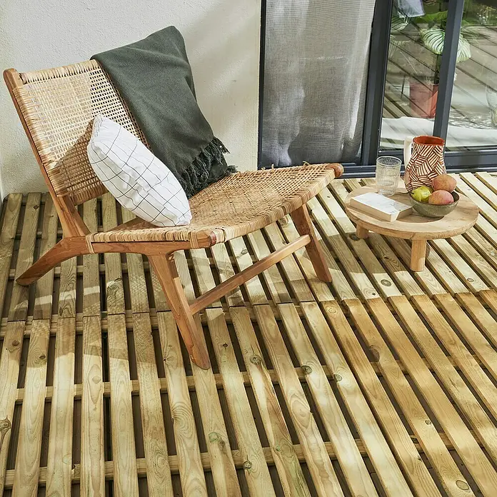 Cerland Loseta de madera Cleia - Pino, 100 x 100 x 3 cm, 1 ud. - Escena acogedora con una silla de ratán en una terraza de madera, adornada con cojines y una manta, junto a una mesa auxiliar con artículos decorativos.