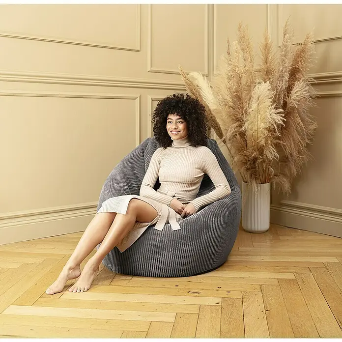 Sitzsack Beanbag Shara - XL, Anthrazit, 95 % Polyester, 5 % Nylon - Eine Frau sitzt entspannt in einem grauen Cord-Sitzsackstuhl, der Komfort und Stil vereint. Perfekt zum Relaxen im Wohnzimmer.