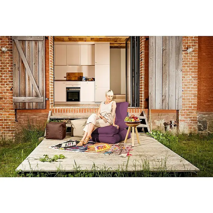 Sitting Point Sitzsack Twist Scuba - 90 x 70 x 80 cm, Aubergine, 100 % Polyester/100 % Polyvinylchlorid - Eine Frau entspannt auf einem lila Sitzsackstuhl im Freien, der auf einem farbenfrohen Teppich auf einer Holzterrasse steht.