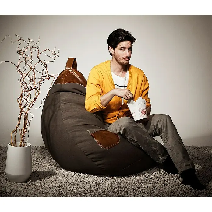 Sitting Point Sitzsack Beanbag Jamie - XL, Braun, 100 % Polyester - Ein Mann sitzt entspannt auf einem braunen Sitzsack mit Lederdetails und isst asiatisches Essen aus einer weißen Box.
