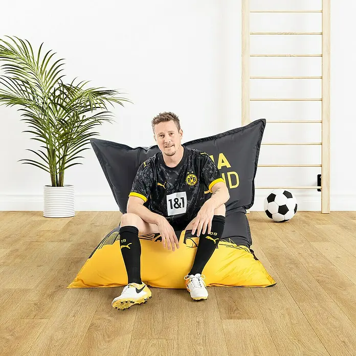 Sitting Point Sitzsack VIP Big Bag - 170 x 130 cm, BVB, 100 % Polyester - Ein Mann sitzt auf einem BVB-Sitzsack, mit Vereinslogo und Sponsorenaufdruck, im Zimmer.