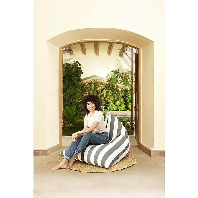 Sitting Point Sitzsack Santorin Calypso - 110 x 115 x 90 cm, Anthrazit/Weiß, 100 % Polypropylen - Eine junge Frau sitzt entspannt auf einem gestreiften Sitzsack im Innenbereich mit Blick auf einen üppigen Garten.