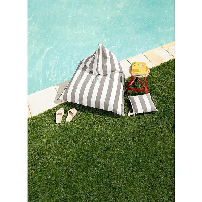 Sitting Point Sitzsack Santorin Calypso - 110 x 115 x 90 cm, Taupe/Weiß, 100 % Polypropylen - Ein gestreifter Sitzsack, ein Kissen, Sandalen, ein Beistelltisch und ein Buch liegen neben einem Schwimmbecken auf Rasen.