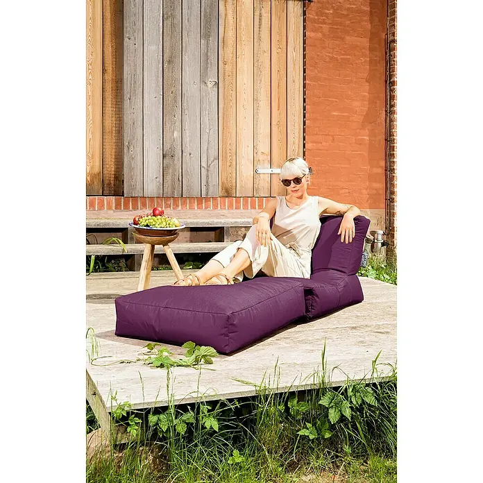 Sitting Point Sitzsack Twist Scuba - 90 x 70 x 80 cm, Aubergine, 100 % Polyester/100 % Polyvinylchlorid - Eine stilvolle Frau entspannt auf einem violetten Lounge-Sessel im Freien, umgeben von Grün und rustikalem Holz.