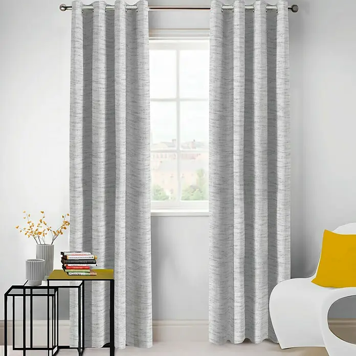 Visillo para ventana Argán - 140 x 250 cm, 100% poliéster, Gris - Un par de cortinas grises con ojales colgadas de una barra de cortina frente a una ventana, añadiendo un toque elegante y moderno a la habitación.