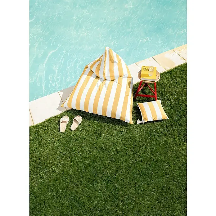 Sitting Point Sitzsack Santorin Calypso - 110 x 115 x 90 cm, Gelb/Weiß, 100 % Polypropylen - Ein gelb-weiß gestreifter Sitzsack und ein Kissen liegen auf dem Rasen neben einem Pool, mit einem kleinen Tisch, einem Buch und Sandalen.