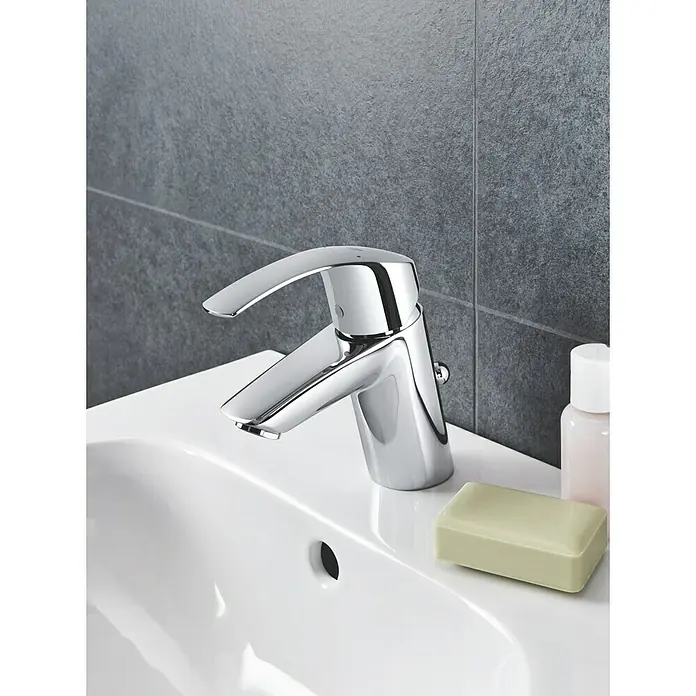 Grohe Eurosmart Waschtischarmatur S-Size - Mit Zugstange, Chrom, Glänzend - Nahaufnahme eines glänzenden Chrom-Wasserhahns auf einem weißen Waschbecken, vor einer grauen, gefliesten Wand.
