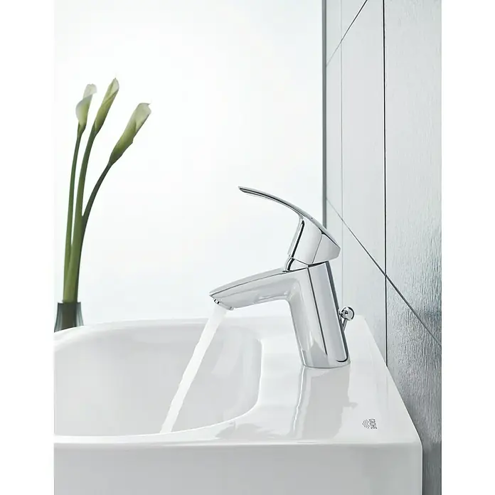 Grohe Eurosmart Waschtischarmatur S-Size - Mit Zugstange, Chrom, Glänzend - Ein glänzender Chrom-Wasserhahn von Grohe auf einem weißen Waschbecken mit fließendem Wasser, akzentuiert durch elegante Blumen und moderne Fliesen.
