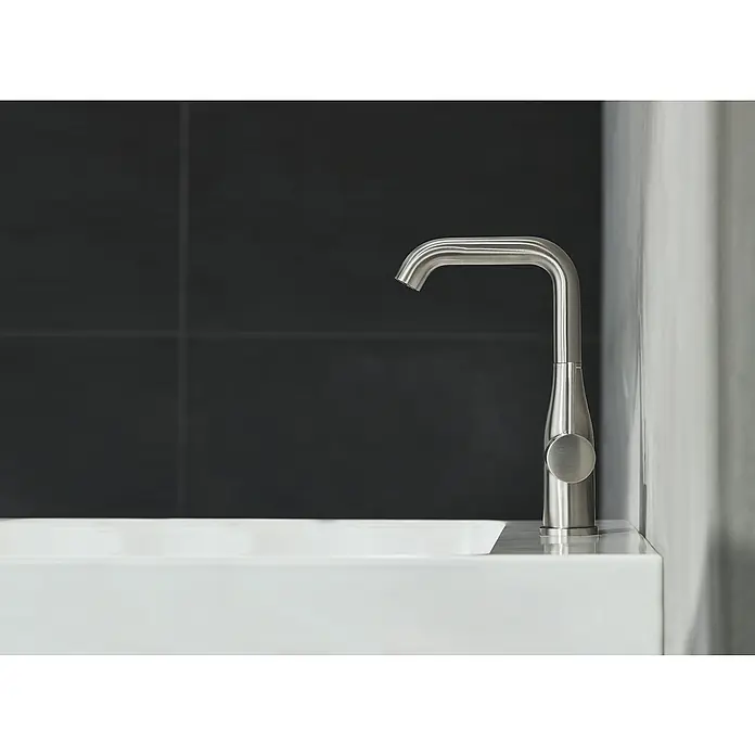 Grohe Essence New Waschtischarmatur L-Size - Mit Zugstange, Supersteel, Glänzend - Moderne Armatur auf einem weißen Waschbecken vor dunklen Fliesen. Die Armatur ist aus gebürstetem Metall.