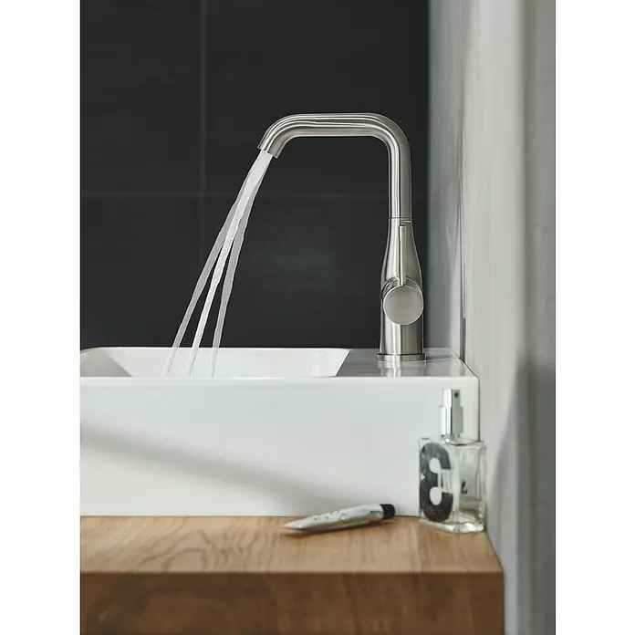 Grohe Essence New Waschtischarmatur L-Size - Mit Zugstange, Supersteel, Glänzend - Nahaufnahme einer modernen Badezimmerarmatur aus Edelstahl mit fließendem Wasser, montiert auf einem weißen Waschbecken mit Holzablage.