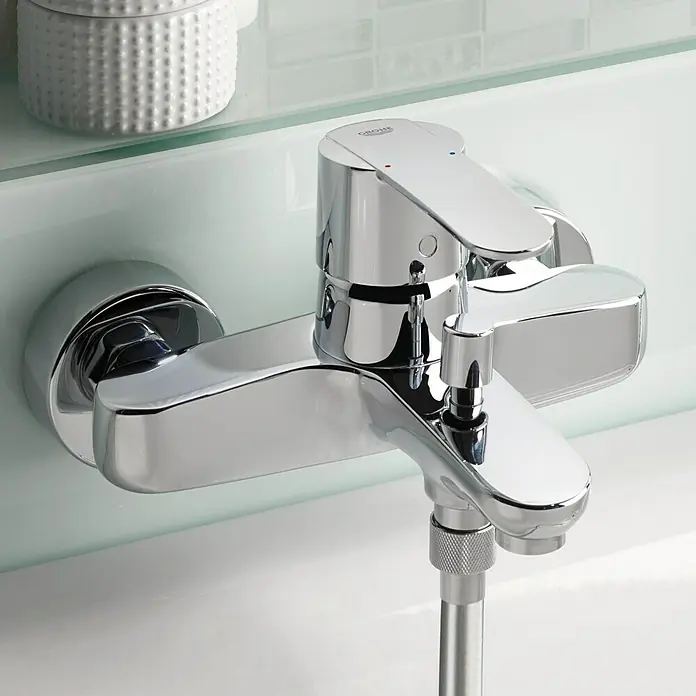 Grohe QuickFix Wave Cosmopolitan Grifo monomando de bañera - Cromo, Resplandeciente - Primer plano de un grifo Grohe brillante montado en la pared de un baño con un recipiente decorativo blanco al fondo.
