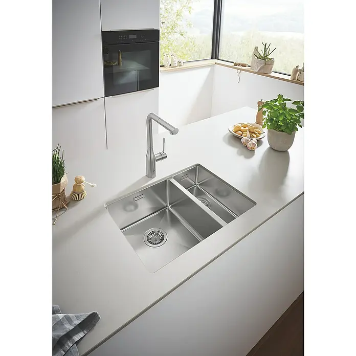 Grohe Essence Spültischarmatur - L-Auslauf, Supersteel, Matt - Doppelspülbecken aus Edelstahl mit modernem Wasserhahn in einer hellen, modernen Küche. Küchengeräte und Pflanzen sind im Hintergrund sichtbar.