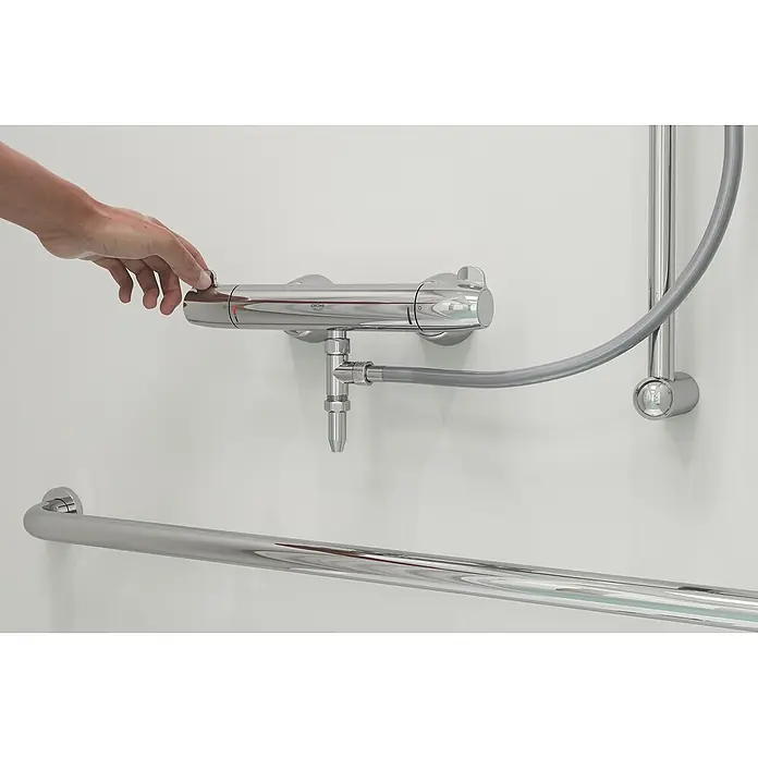 Grohe T-Stück Universal - Passend für: Wannen- und Brausearmaturen, Gewinde: ½″ - Detailaufnahme einer Hand, die einen Grohe Duschmischer bedient, mit Fokus auf Armatur, Schlauch und Haltegriff an einer weißen Wand.