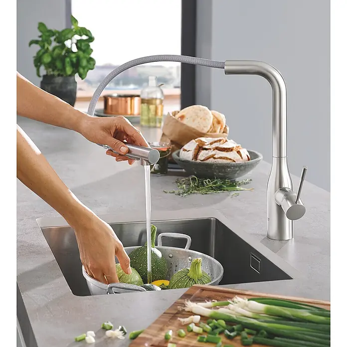 Grohe Essence Spültischarmatur - L-Auslauf, Supersteel, Matt - Eine Person wäscht Gemüse unter einem modernen Küchenarmatur mit Ausziehbrause in einer Edelstahlspüle.