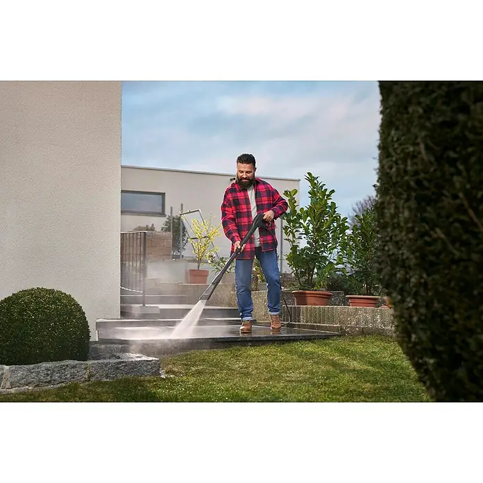 Kärcher Strahlrohr eco!Booster 130 - Passend für: Kärcher Hochdruckreiniger der Klassen K4, 615 x 108 x 51 mm - Ein Mann im Garten reinigt mit einem Kärcher Hochdruckreiniger eine Steinterrasse, umgeben von grünen Pflanzen und Büschen.