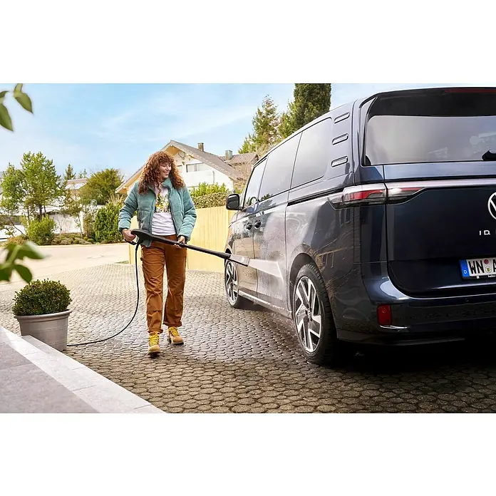 Kärcher Strahlrohr eco!Booster 130 - Passend für: Kärcher Hochdruckreiniger der Klassen K4, 615 x 108 x 51 mm - Eine Frau reinigt einen VW ID. Buzz mit einem Hochdruckreiniger auf einer gepflasterten Auffahrt vor einem Haus.