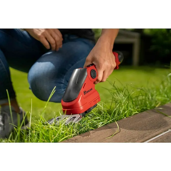 Hurricane Akku-Gras- & Strauchschere HGS-I-120 - 12 V, Li-Ionen, 1 Akku - Eine Person benutzt eine rote Hurricane-Gartenschere, um Gras entlang eines Holzdecks zu schneiden.