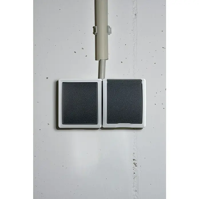 Aqua Form Feuchtraum-Schalterkombination - Anthrazit, Ausrichtung Schalterprogramm: Horizontal, Aufputzmontage, IP44 - Doppelte Lichtschalter mit weißem Rahmen und schwarzen Knöpfen an einer weißen Wand montiert, mit Kabelkanal darüber.