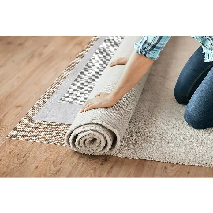 D-c-fix Teppichgleitschutz Loom - 180 x 120 cm - Eine Person rollt einen grauen Teppich auf einem Holzboden mit einer Teppichunterlage aus, um ein Verrutschen zu verhindern.
